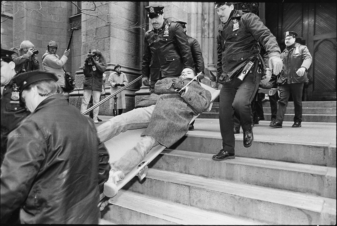 Black and white image of four policeman carrying male protestor down cathedral steps on stretcher.