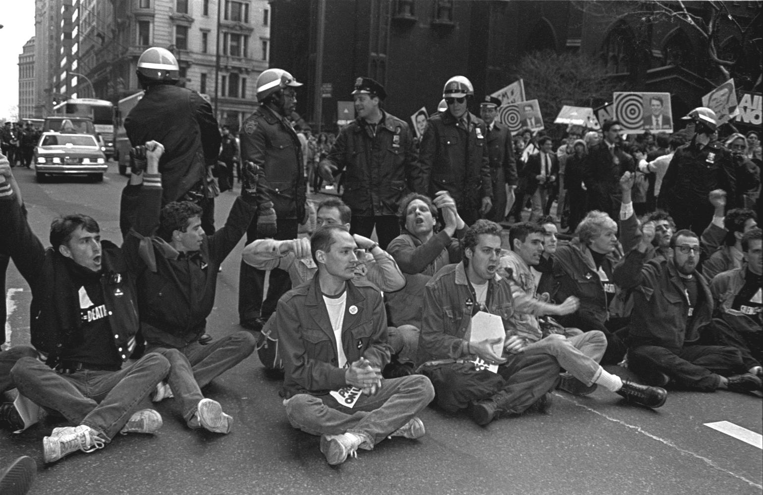 Black and white photo of demonstrators blocking street by sitting while a line of police stand behind them.