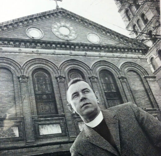 Black and white photo of man in front of church wearing a minister's shirt and jacket.