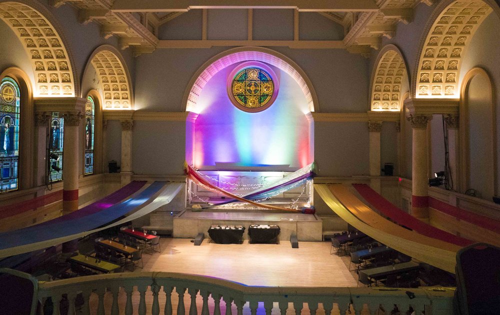 Interior of church decorated with rainbow colored lighting and rainbow cloth stretched above tables set up for a meal.