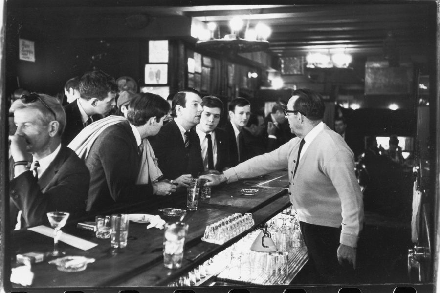 Black and white image of a group of approximately 12 white men in suits and ties being served cocktails at a bar. Wood beams line the ceiling with wagon wheel style pendant fixtures.