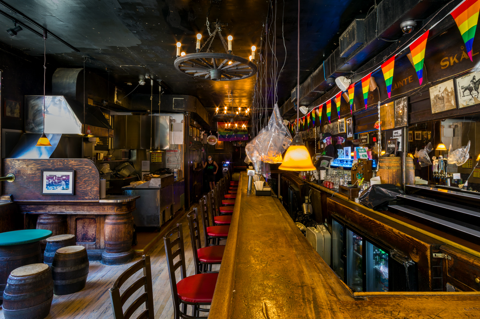 A wooden bar stretches the length of the room with red barstools. Green round tables are surrounded by barrels used as stools. A stream of rainbow pendants hang above the bar.