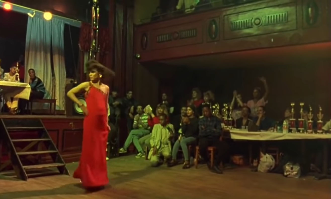 The room is dark wood with some stage lighting. A black performer in red dress has one hand on hip as they look to the side. People surround the performer as an audience. They are a diverse crowd. Trophies sit upon a table with a white tablecloth.