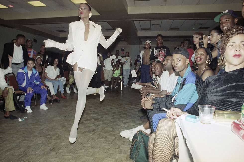 A crowd of primarily people of color watch a black performer in white stockings and a white blazer performer in the middle of the room.