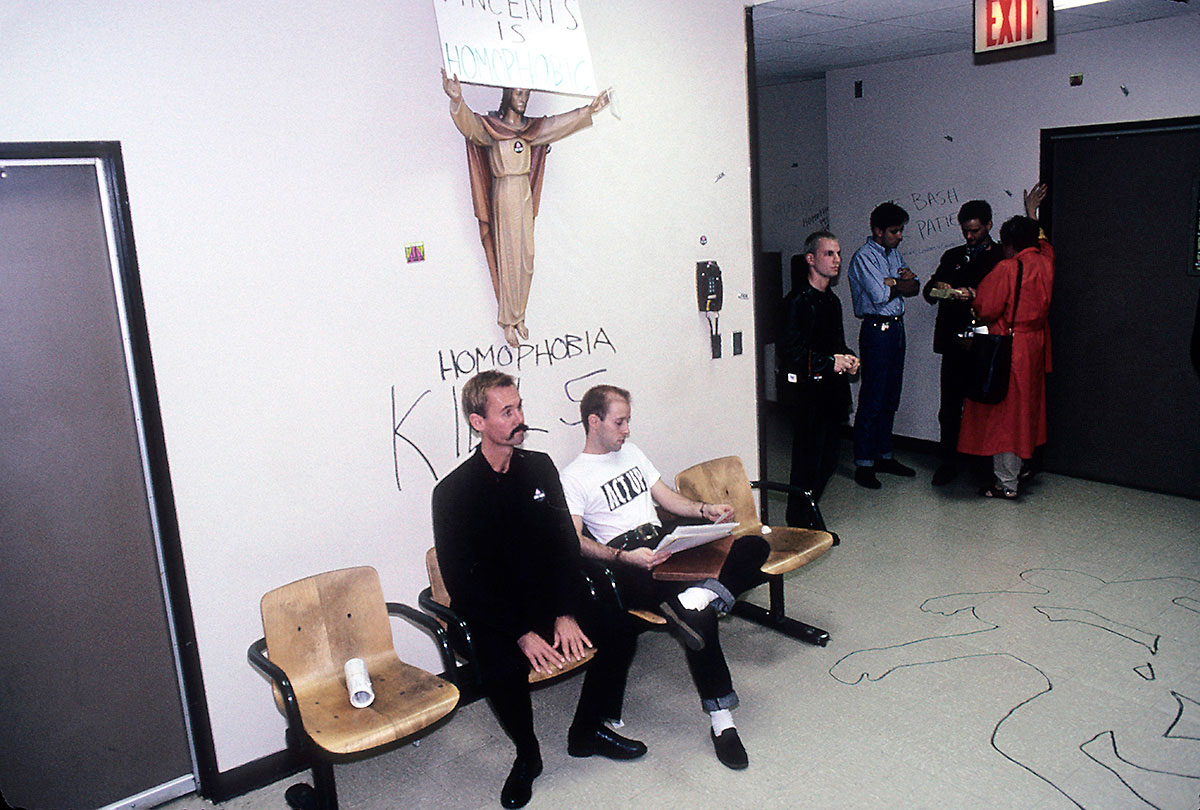 Two men sitting in waiting room under Jesus with graffiti on wall that says Homophobia Kills