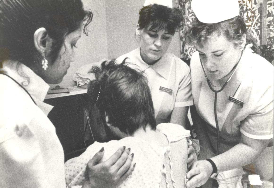 Black and white photo of three women in nurse uniform supporting a male patient with back turned to camera