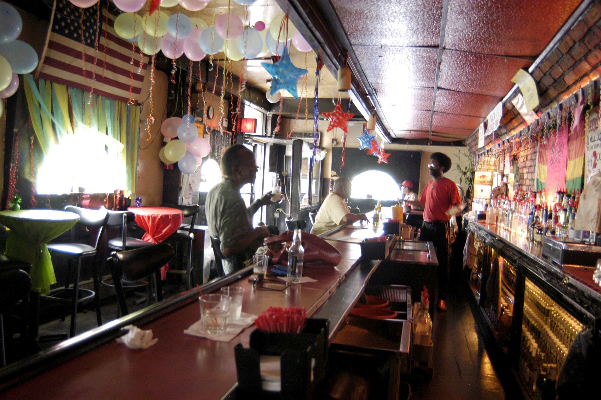 A bar with three customres and a bartender in red shirt with afro hairstyle. Colorful balloons fill the ceiling. Read acoustic tile is above the bar area. Daylight is seen through the windows.