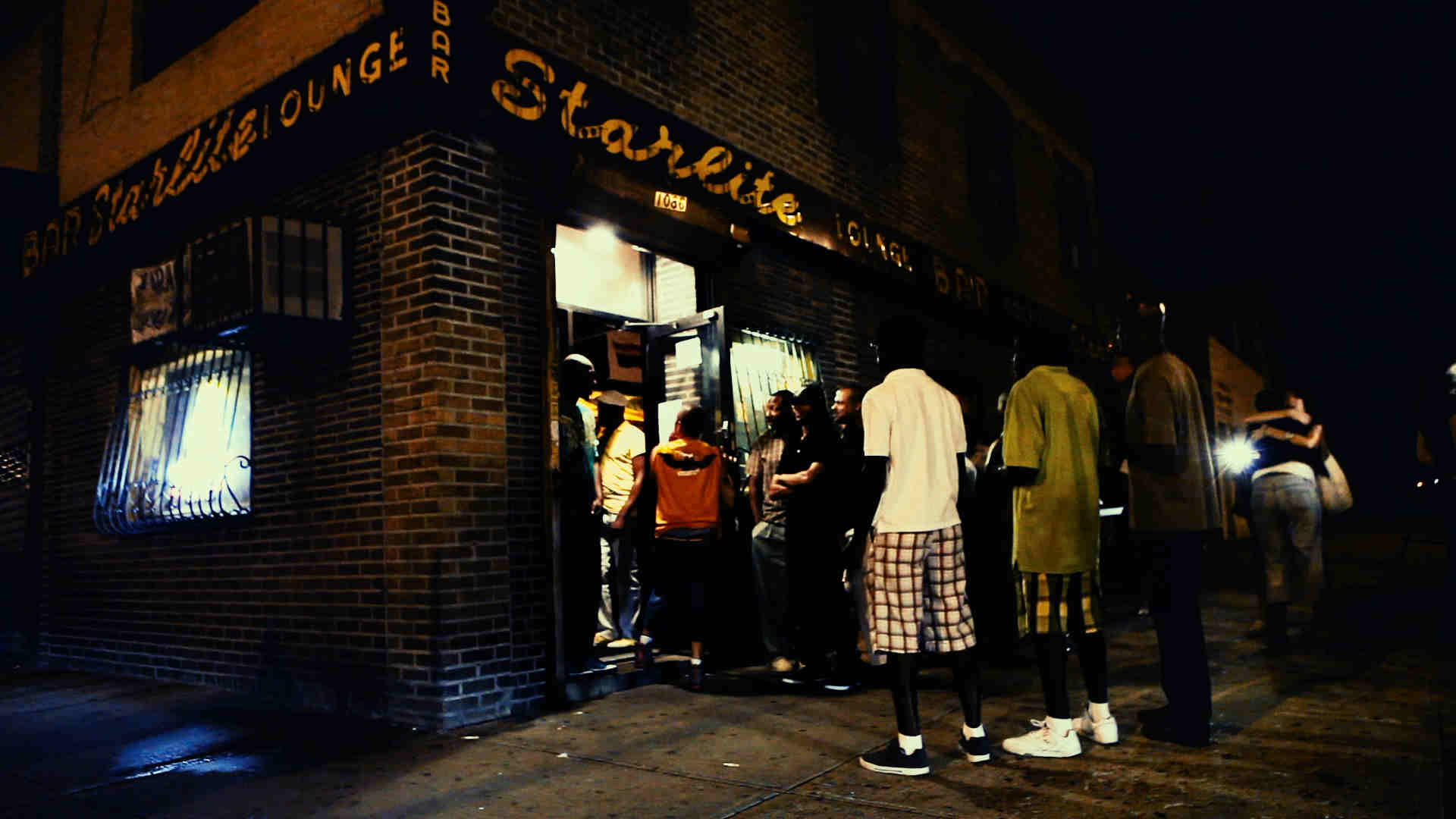 Night time exterior of bar with people lining up on sidewalk to enter.
