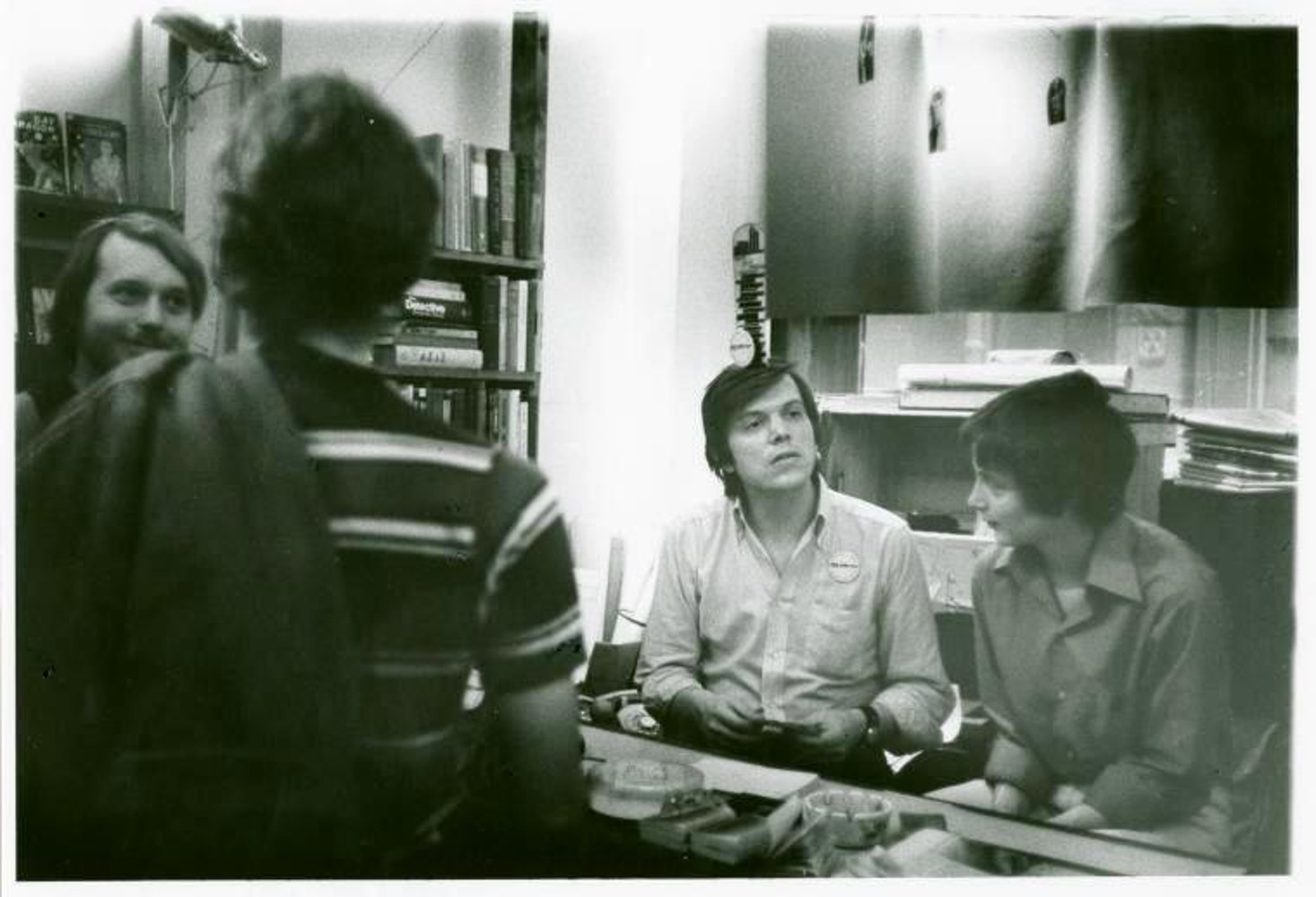Black and white image of four people. Two seated and two standing. Bookshelves are on the left side of the image.