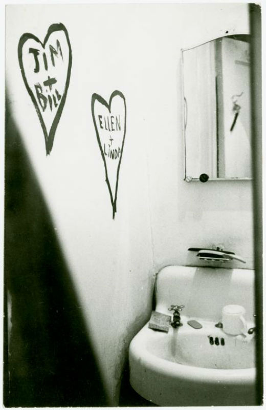 Black and white image of small bathroom with sink and mirror. Two hearts are drawn in the wall in thick marker inside one says, 