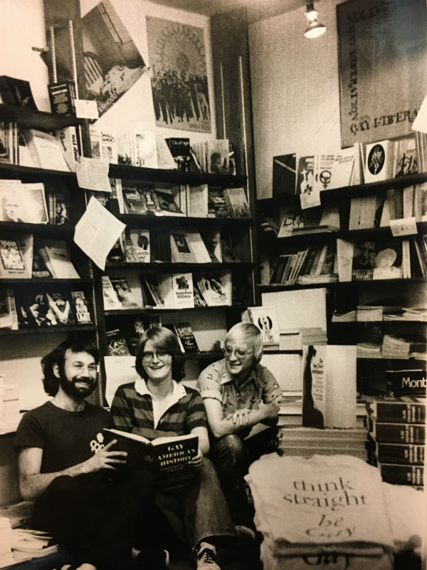 Black and white image of three people sitting on floor reading a book. They are in a room full of bookshelves.