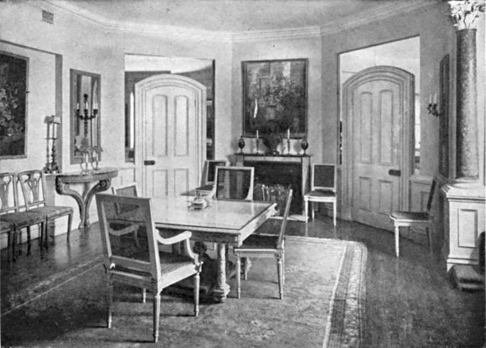 Black and white image of a dining room. There are two angled walls with arched doorways surrounded by mirrors.