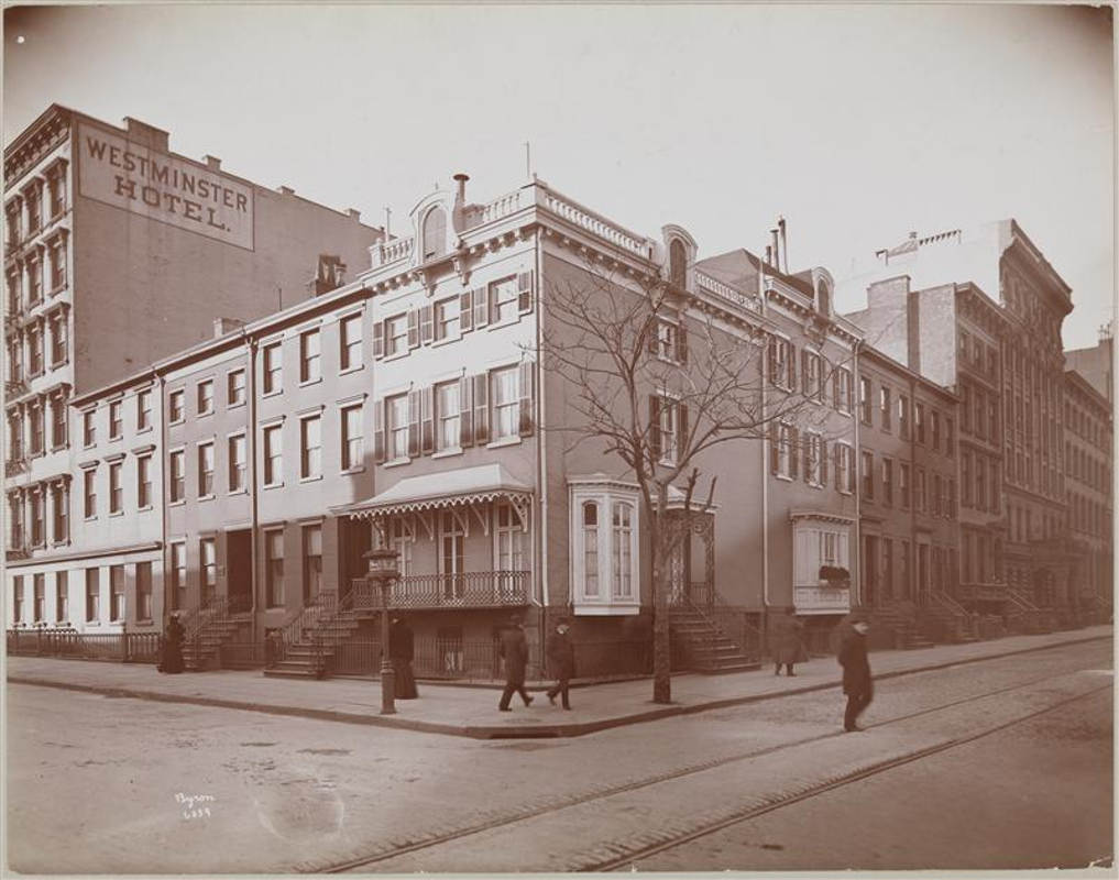 Sepia toned image of townhouse in NYC from 1900. It is winder with no leaves on the trees. The building at corner has awning and ornamental parapet.