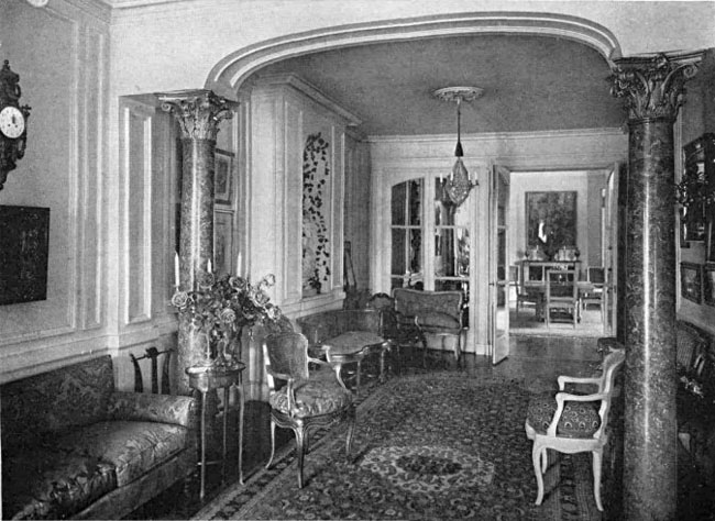 Black and white photo showing a room with two marble corinthian columns and an arch. Floral patterned upholstry on sofa and chairs.