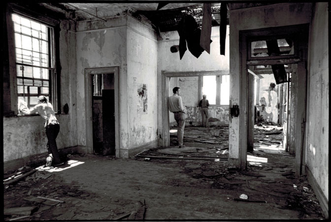 Black and white photograph of the interior of an abandoned building. The ceiling is falling and the walls peeling. Three men are in the building all in different spots not facing each other.