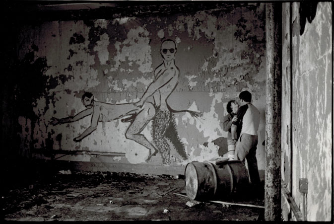 Black and white photograph of the interior of an abandoned building. Graffiti on the wall shows two men engaged in sex. Two men are being intimate behind an overturned barrel in the corner of the room.