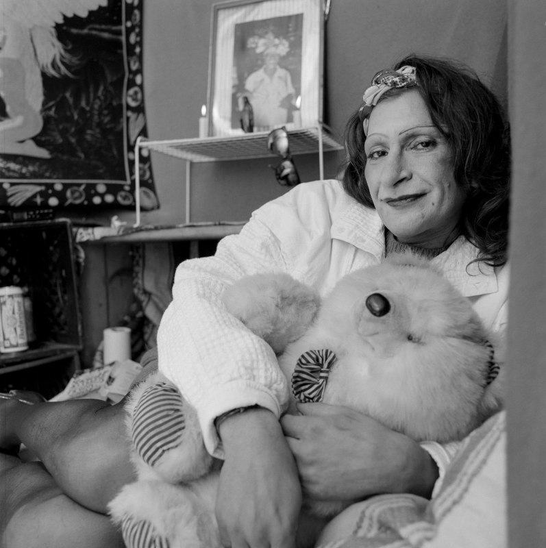 Black and white photo of a person in robe holding a stuffed teddy bear. Behind her is a photo with two candles burning next to it.