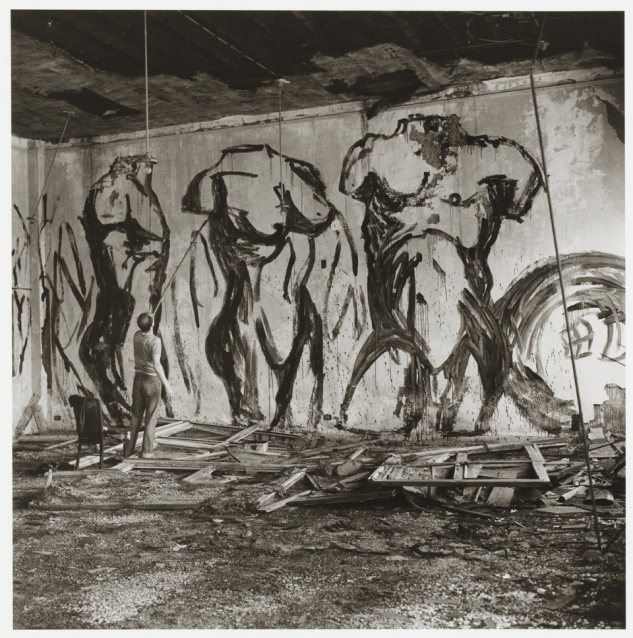 Black and white photo of a man painting a mural on a wall in an abandoned building. He is painting two male and one female torso with black outlines