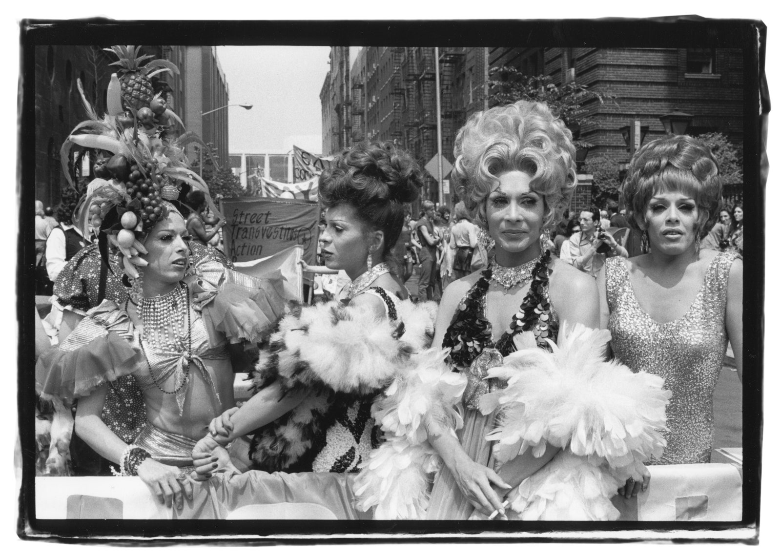 Black and white photograph of four drag queens in the middle of the street. Two face each other while the others look forward beyond the camera frame