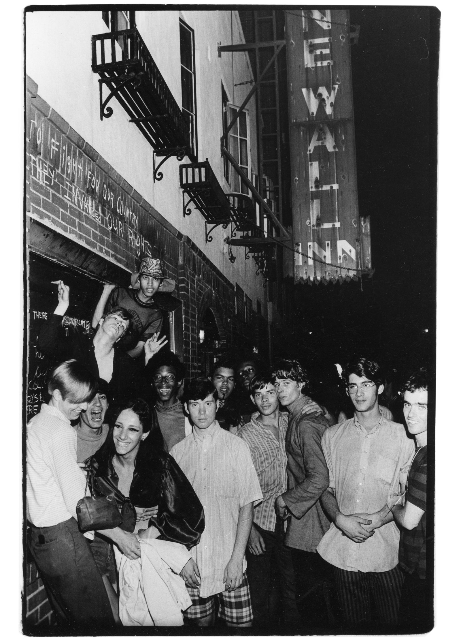 Black and white photo of people outside of Stonewall Inn. People are crowded on sidewalk and two people hang out from bar window.