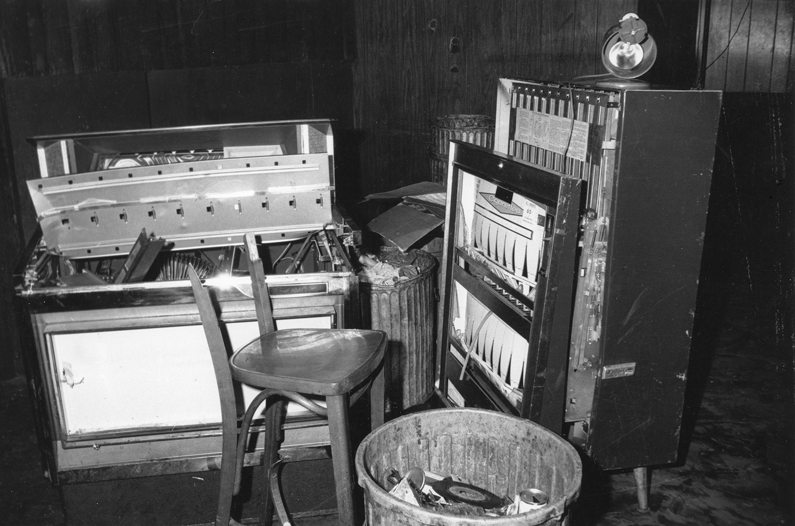 Black and white photo showing a damaged juke box, barstool, and cigarette machine.