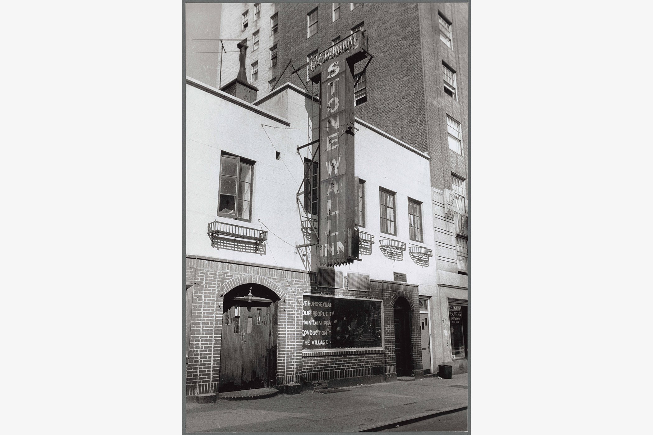 Black and white image of the exterior of the Stonewall Inn. The building is two stories with brick on the first floor, a large wood double door, and white painted plaster second floor exterior.