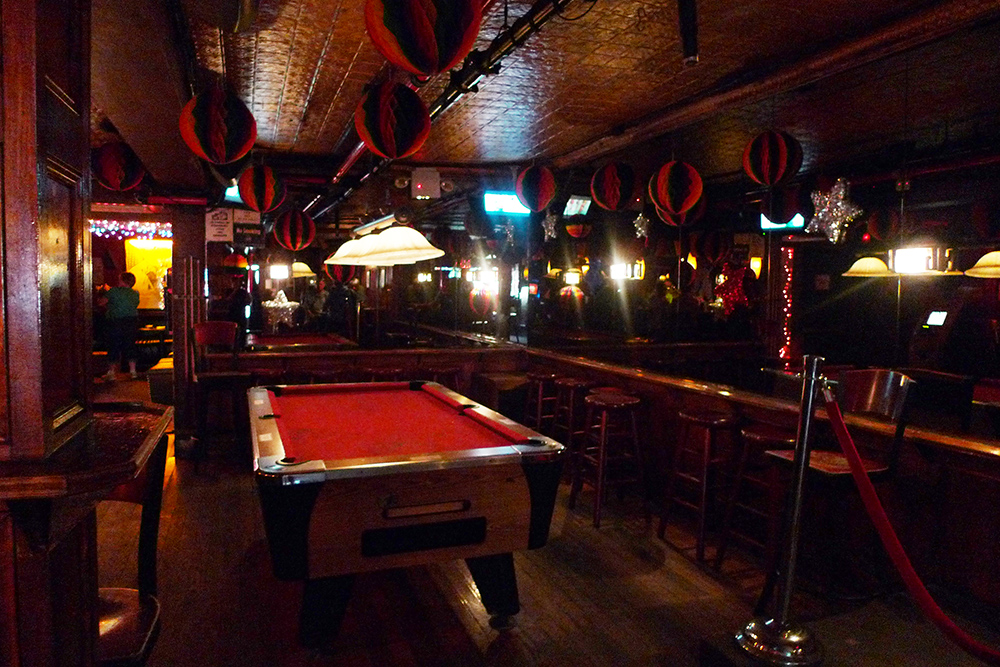color image showing pool table in small room. barstools surrond the edge of the room with mirrored walls above. Round rainbow decorations hang from a painted tin ceiling.
